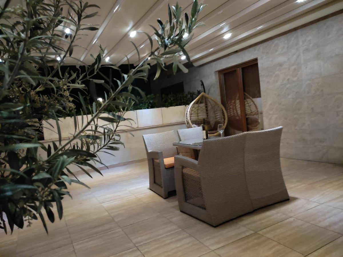 A covered terrace features a set of wicker armchairs and a matching table, creating a cozy seating area. Soft lighting enhances the ambiance, while a decorative plant adds a touch of greenery. The stone walls provide a serene backdrop to the space.