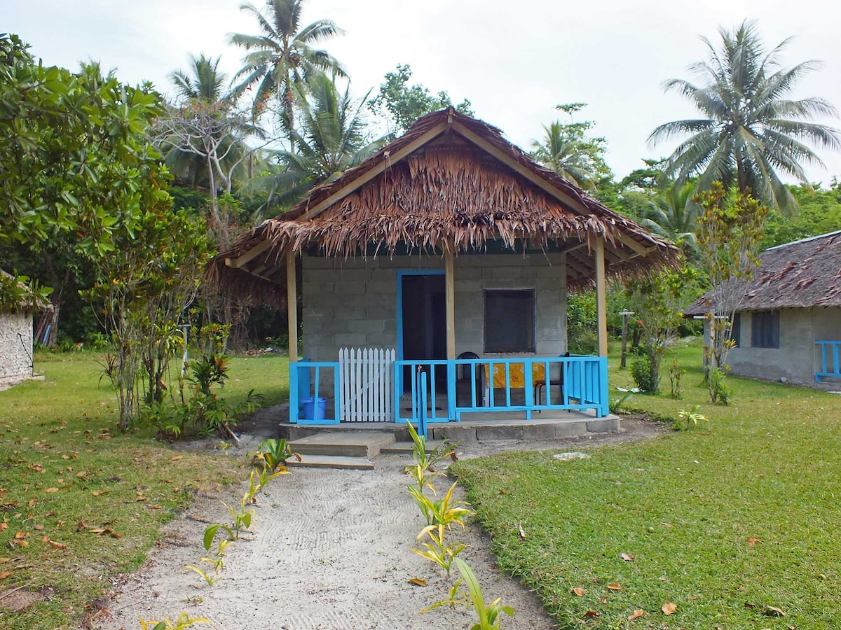 The bungalow features a traditional Natangura thatch roof and a front porch with blue trim. Surrounded by greenery, the structure is set in an open area with palm trees in the background. A path lined with plants leads to the entrance.