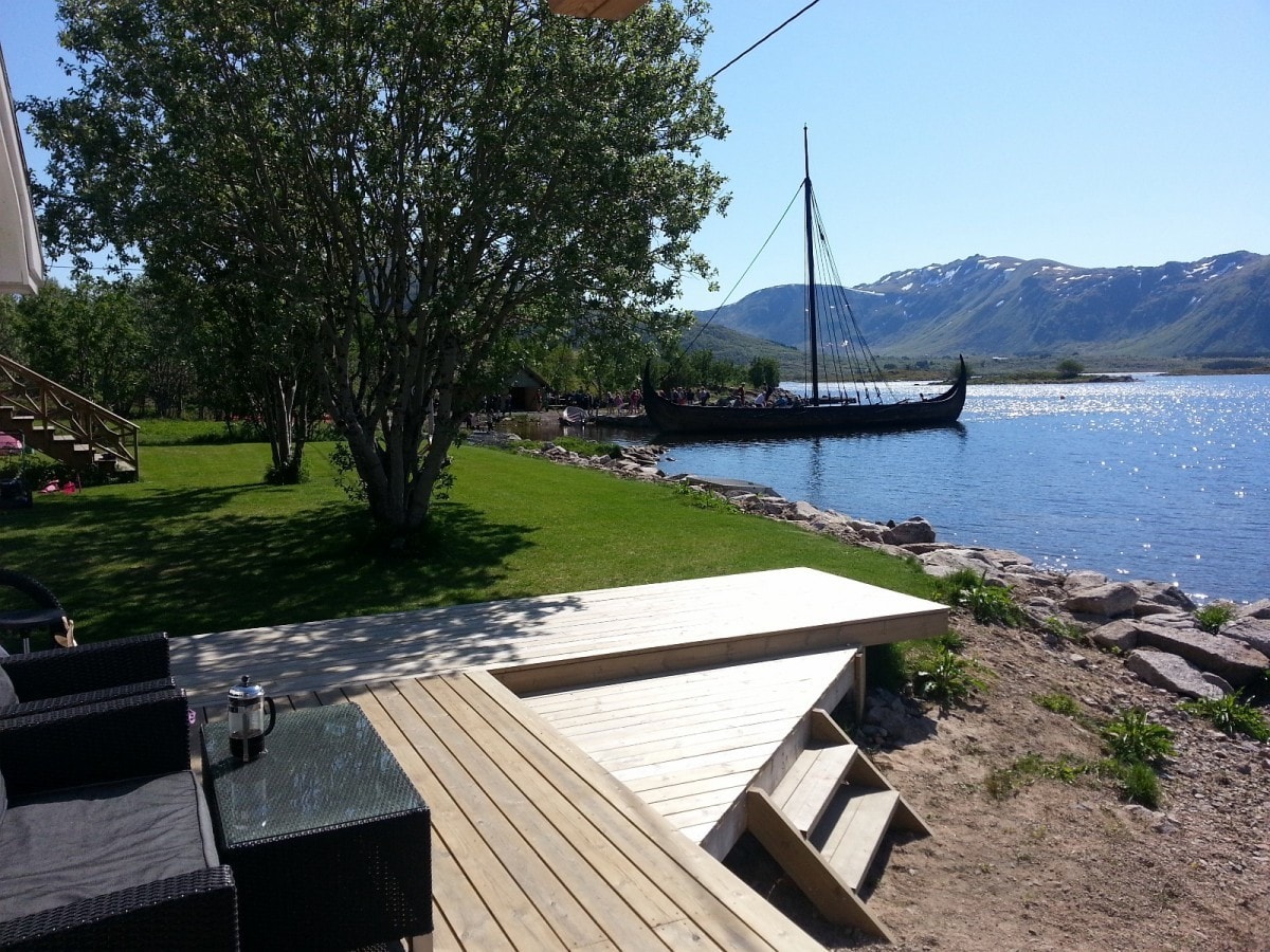 A wooden deck extends towards the water, framed by manicured grass and a small tree. In the background, a traditional boat rests by the shore, with mountains visible across the calm sea under a clear blue sky.