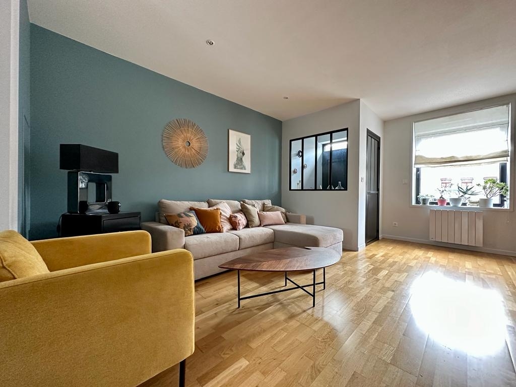 A spacious living area features a light-colored sofa adorned with various cushions, complemented by a mustard yellow armchair. A round coffee table is positioned centrally, with natural light flowing in from a nearby window, creating an inviting and bright environment.