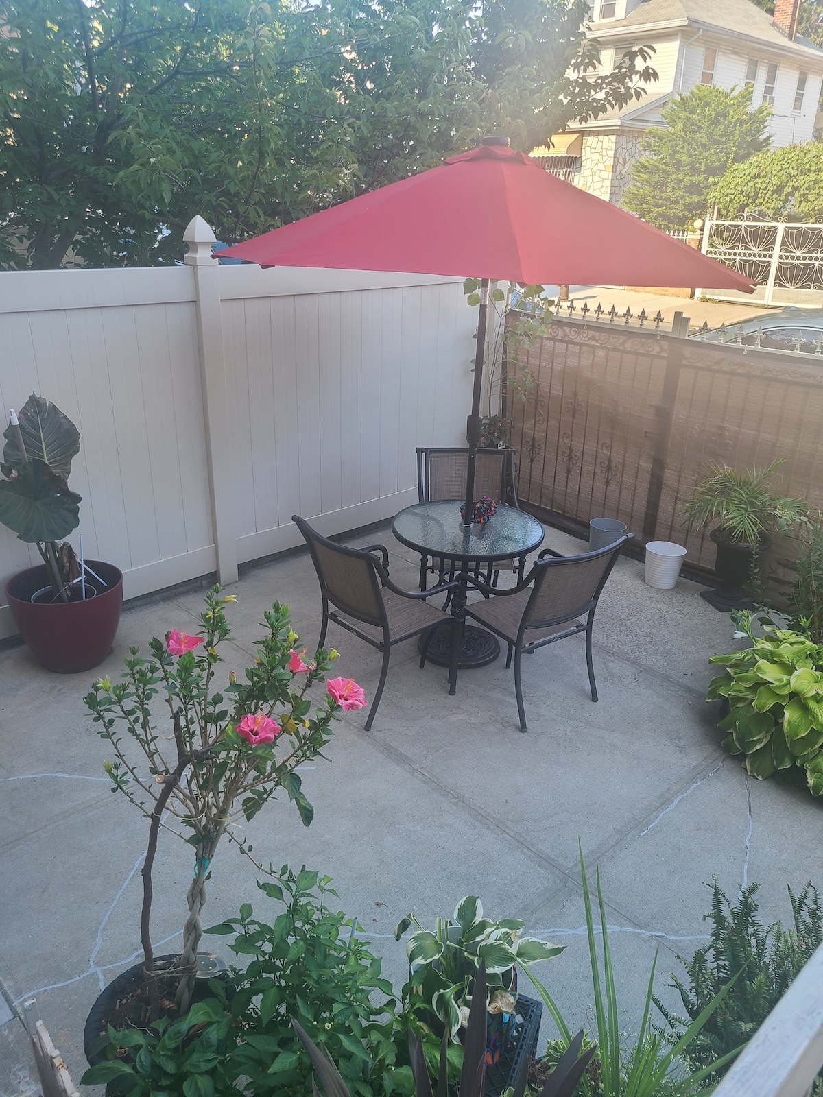 A quaint outdoor patio features a round glass table surrounded by four metal chairs and a large red umbrella providing shade. Potted plants, including vibrant flowers and greenery, enhance the space. A fenced boundary offers privacy while allowing sunlight to filter through.