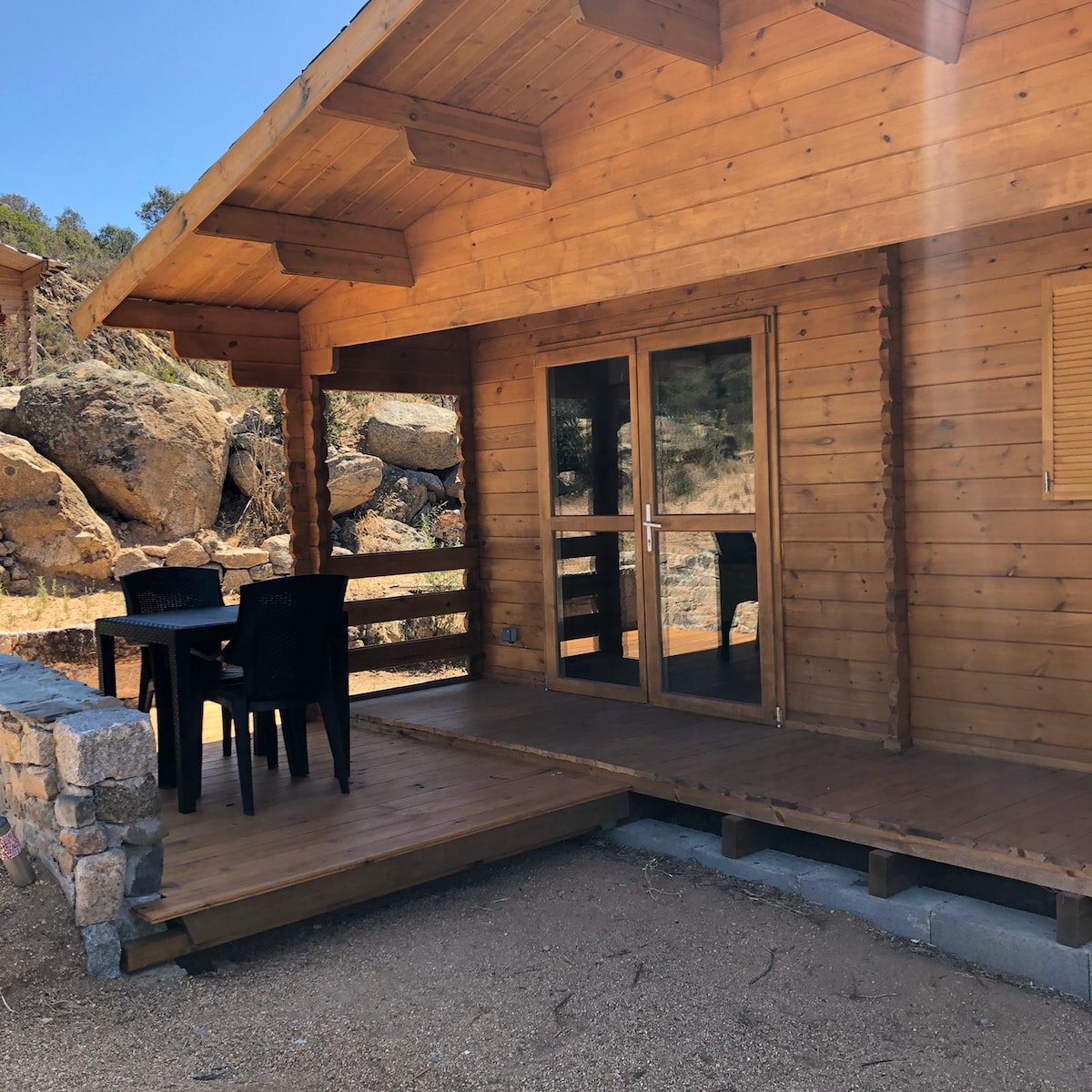 A wooden chalet exterior is displayed, featuring a covered porch with a small table and four chairs. The entrance is framed by large glass doors, allowing for natural light to flow in. Surrounding rocky terrain adds to the serene setting.