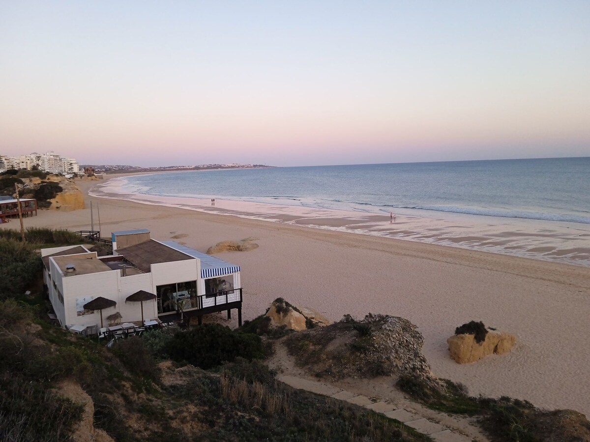 A sandy beach extends along the shoreline, meeting gentle waves that reflect soft colors of the sky. A low white building with a striped awning is visible, accompanied by several umbrellas and seating areas. The tranquil atmosphere provides a sense of relaxation and closeness to nature.