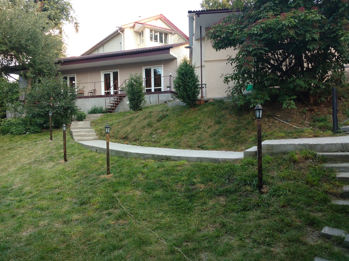 A landscaped garden path leads to the studio, flanked by neat grass and small lanterns. The building features a mix of light and dark exteriors, with large windows that invite natural light. Surrounding greenery adds to the serene environment.