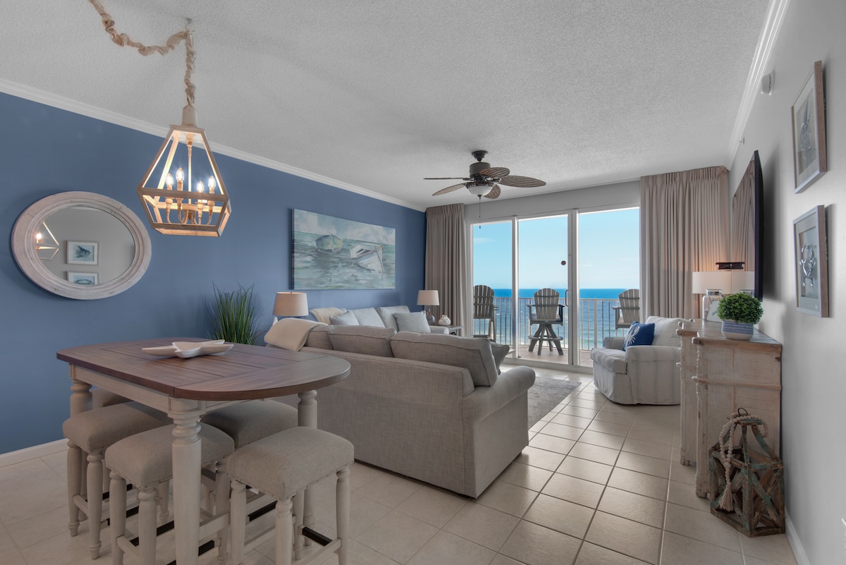 The living area features a spacious layout with a comfortable sofa and several accent chairs. A dining table with four stools is positioned nearby. Large sliding glass doors allow natural light and showcase views of the gulf, complemented by soft blue wall tones and beach-themed decor.