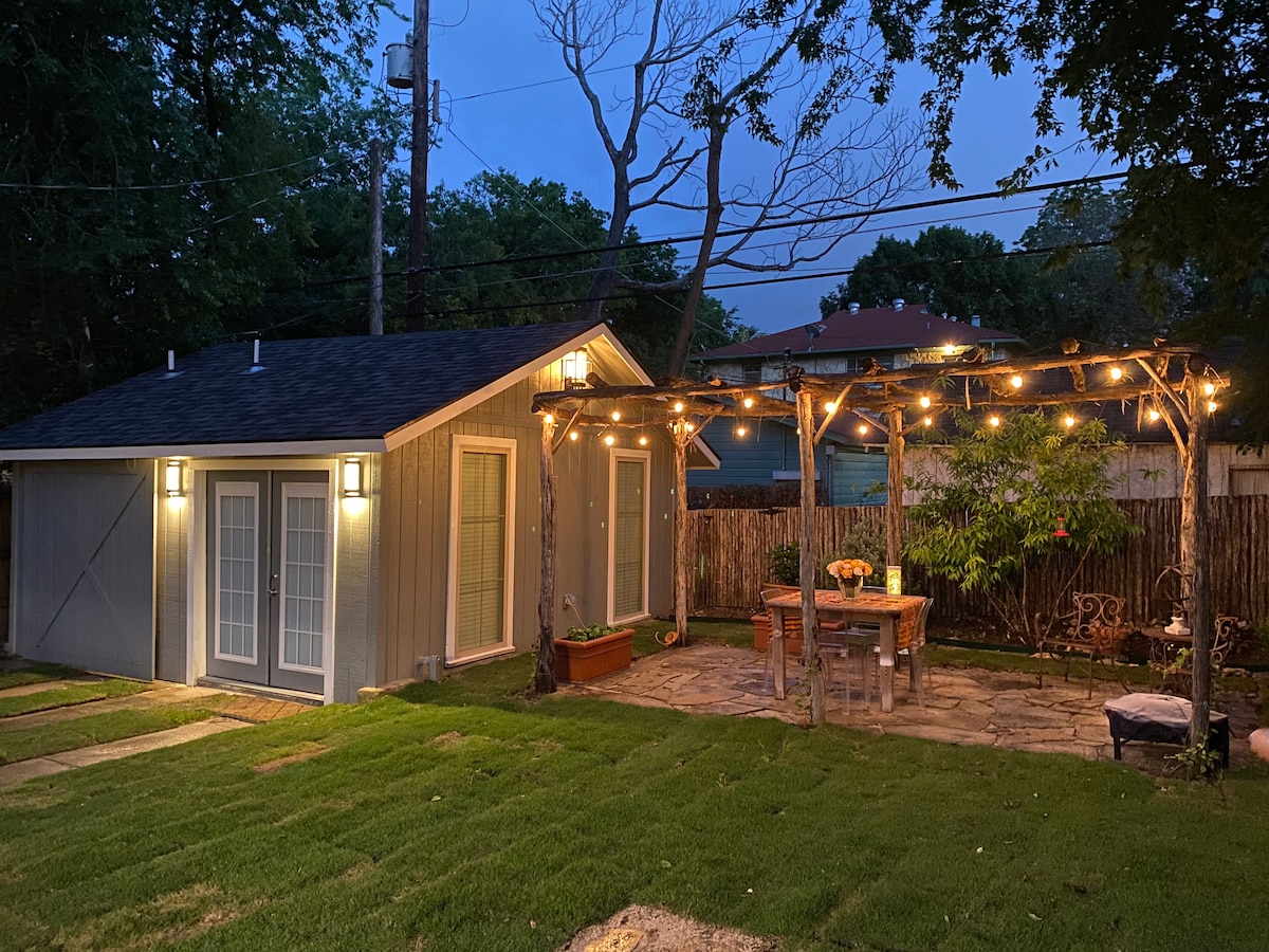 The exterior of the cottage is illuminated by string lights overhead, creating a warm ambiance. A stone patio is visible in front of the cottage, furnished with a wooden table and chairs. Lush green grass surrounds the area, with trees adding natural elements.