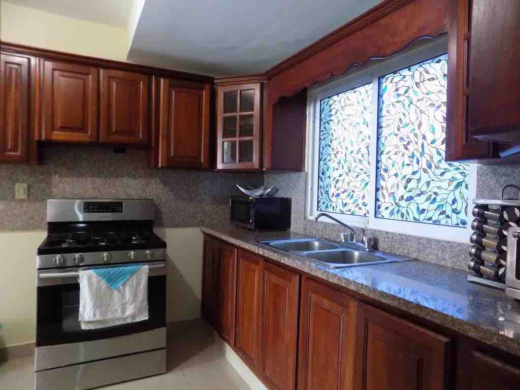 A kitchen area features dark wooden cabinetry and a granite countertop. An oven is placed alongside a microwave and a two-basin sink. Natural light enters through a window adorned with decorative glass, enhancing the space's functionality.