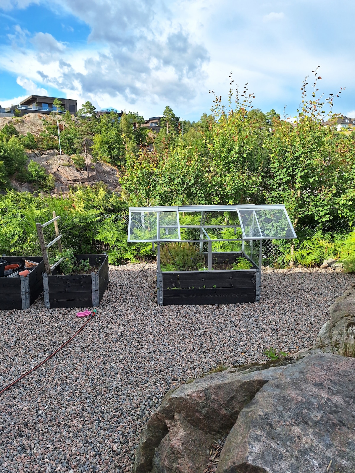 A garden area features raised planter boxes filled with various plants. A greenhouse is positioned next to the boxes, allowing for sunlight exposure. Surrounding greenery includes lush trees and shrubs, creating a natural setting. Gravel paths provide easy access through the garden.