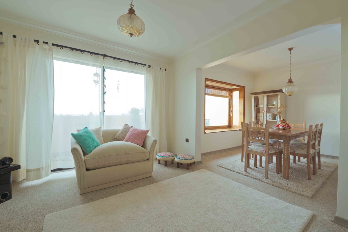 A bright and airy living area features large windows draped with sheer curtains, allowing natural light to fill the space. A comfortable sofa with colorful cushions sits beside a textured area rug. Adjacent, a dining area is highlighted by a wooden table surrounded by six chairs, with a cabinet displaying decorative items.