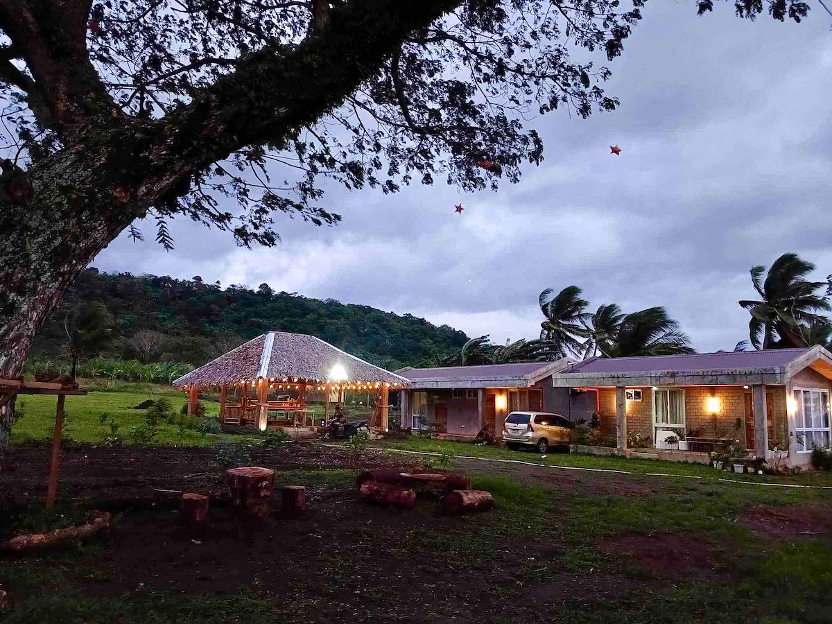 A serene outdoor view features two cabanas situated under a large tree, illuminated by soft string lights. The natural landscape reveals hills in the background, while scattered logs create a rustic seating area on the grassy ground.