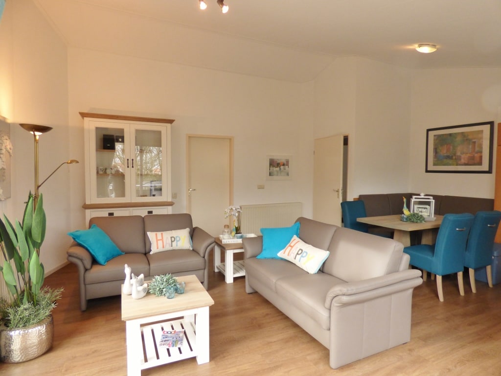 A spacious living area is furnished with light gray sofas, featuring decorative cushions. A coffee table is positioned at the center. A dining area with blue chairs and a wooden table is visible in the background, complemented by warm wooden flooring.