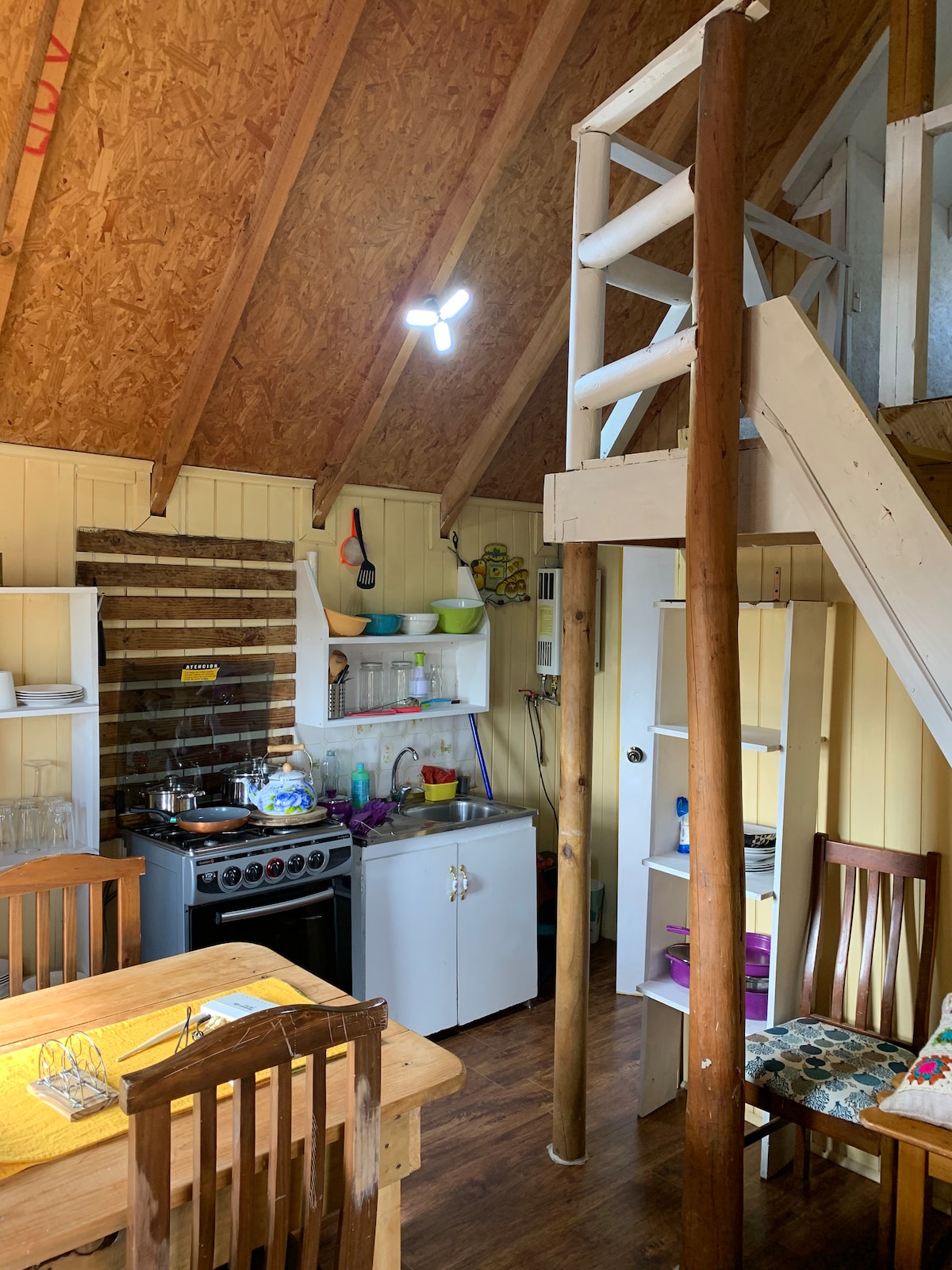 An open kitchen space features essential appliances, including a stove and refrigerator, along with organized shelves holding various kitchenware. A wooden table with chairs occupies the center of the area, while a spiral staircase leads to a lofted sleeping area, enhancing the layout's functionality.