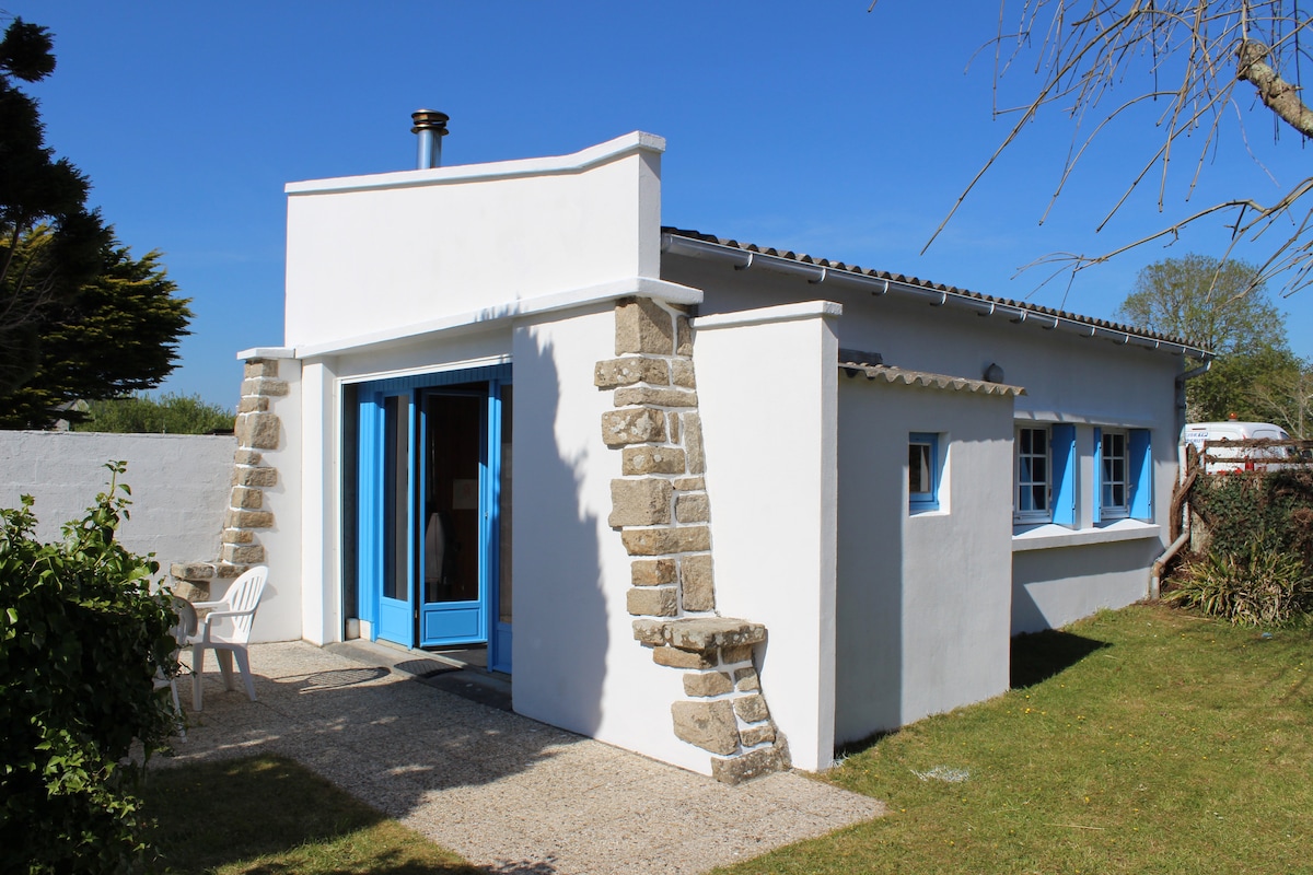 A charming white cottage is shown with blue window frames and contrasting stone accents. A small patio area is visible, equipped with a white chair, and a well-maintained lawn surrounds the property. Clear skies above create a bright atmosphere.