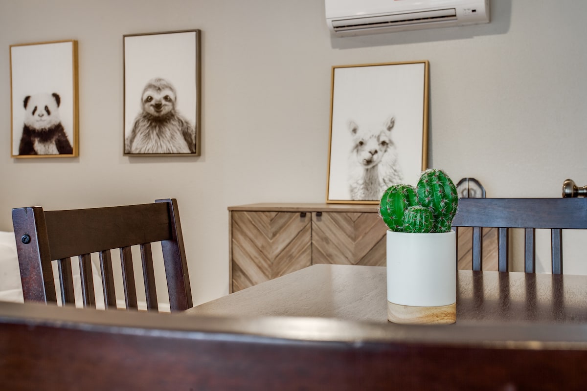 The dining area features a wooden table set against a neutral wall. A small potted cactus serves as the centerpiece, complemented by framed animal portraits in the background. The space is designed with a modern, cozy aesthetic, with light wooden accents visible in the furniture.