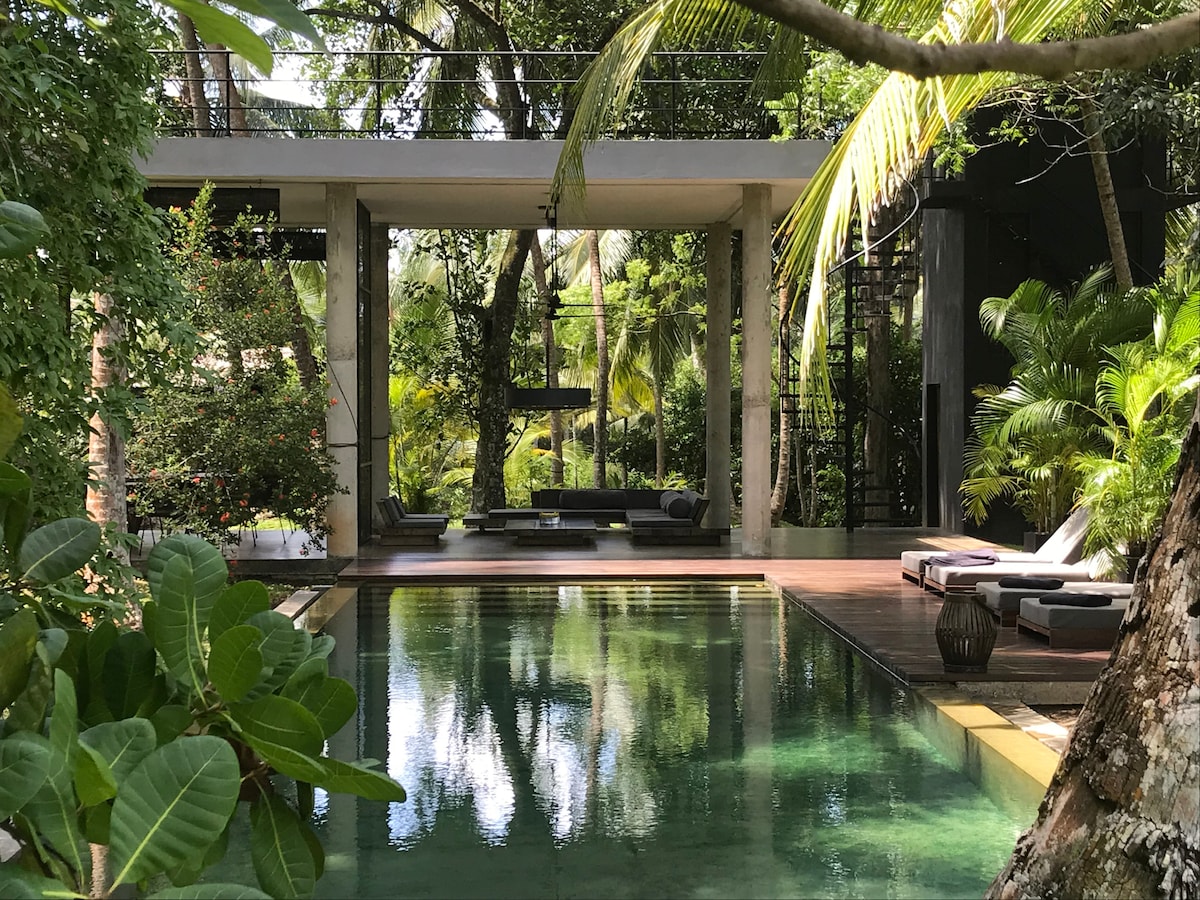 A serene saltwater swimming pool is surrounded by lush greenery, with wooden decking offering seating areas. The design incorporates open spaces that invite natural light, while tropical plants frame the scene, enhancing the overall tranquility of the eco-resort.