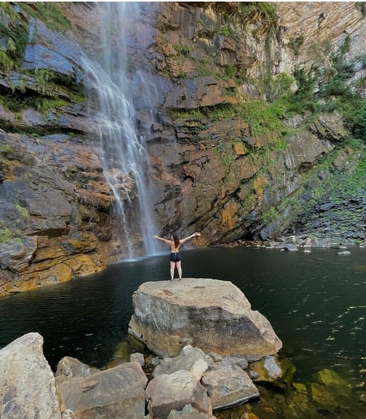 A person stands with arms outstretched on a large rock by a serene pool, surrounded by towering cliffs. A flowing waterfall cascades down the rocky face, adding a sense of tranquility to the natural landscape.