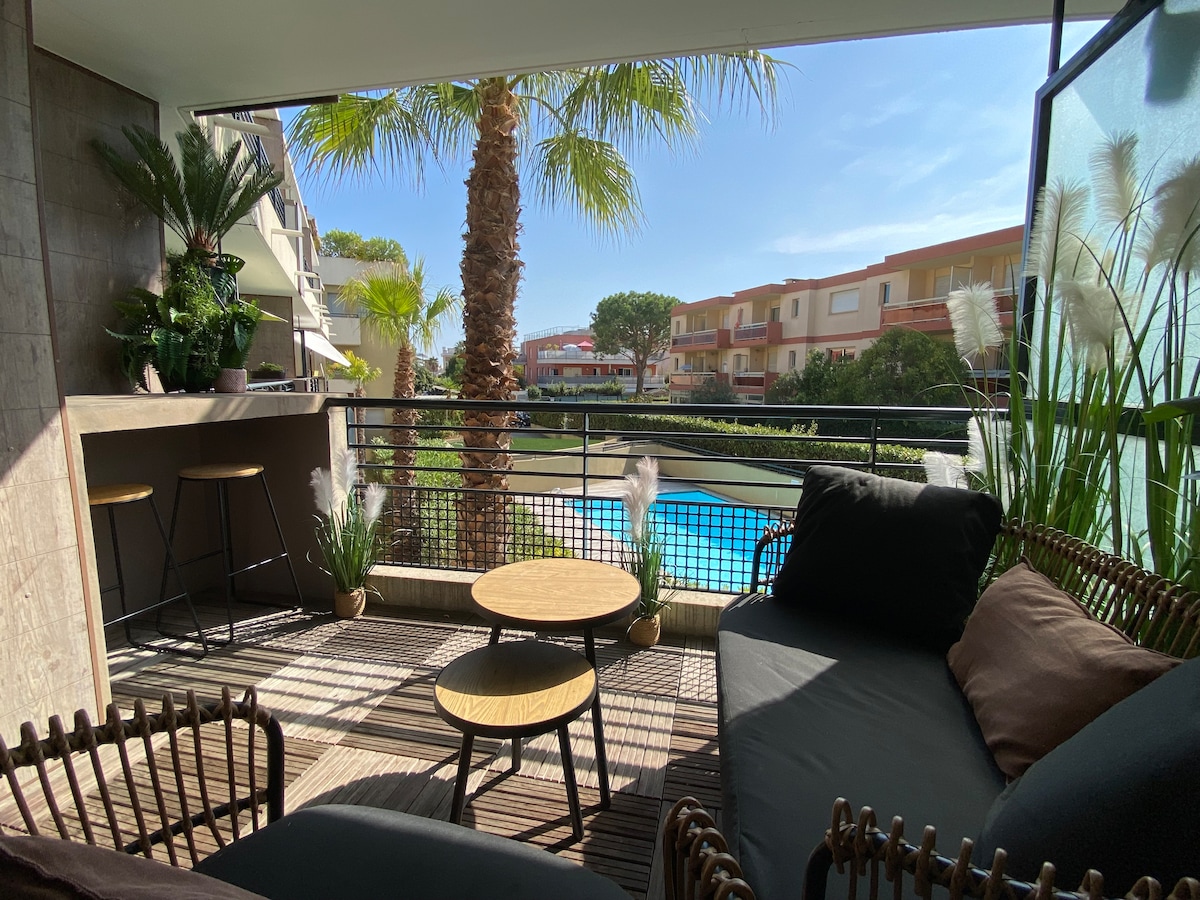 A private terrace features comfortable seating arrangements and small, round tables. Lush greenery surrounds the area, including palm trees visible in the background. A sparkling blue pool can be seen just beyond the railing, with sunlight illuminating the outdoor space.