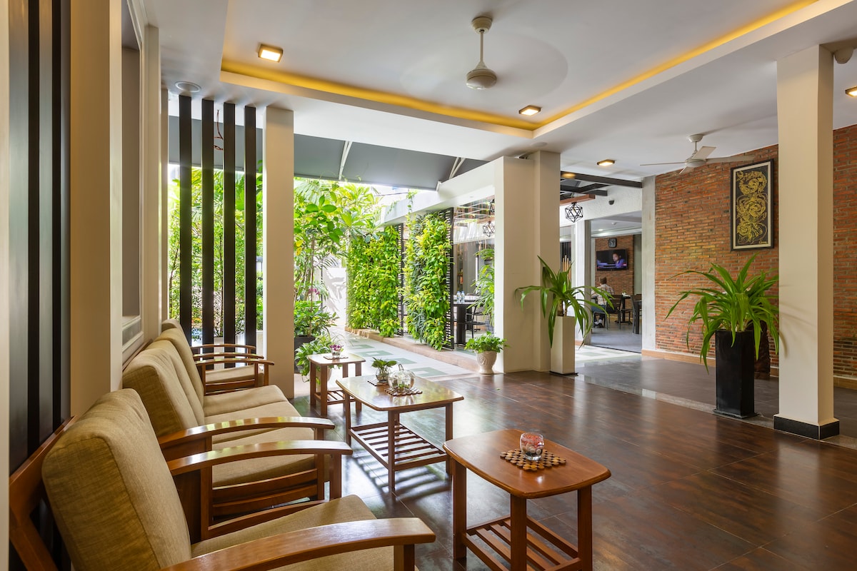 An open and airy common area features wooden seating with soft cushions, complemented by a coffee table. Lush greenery is visible through large windows, creating a connection to the outdoors. Smooth wooden floors and soft lighting enhance the space's welcoming ambiance.