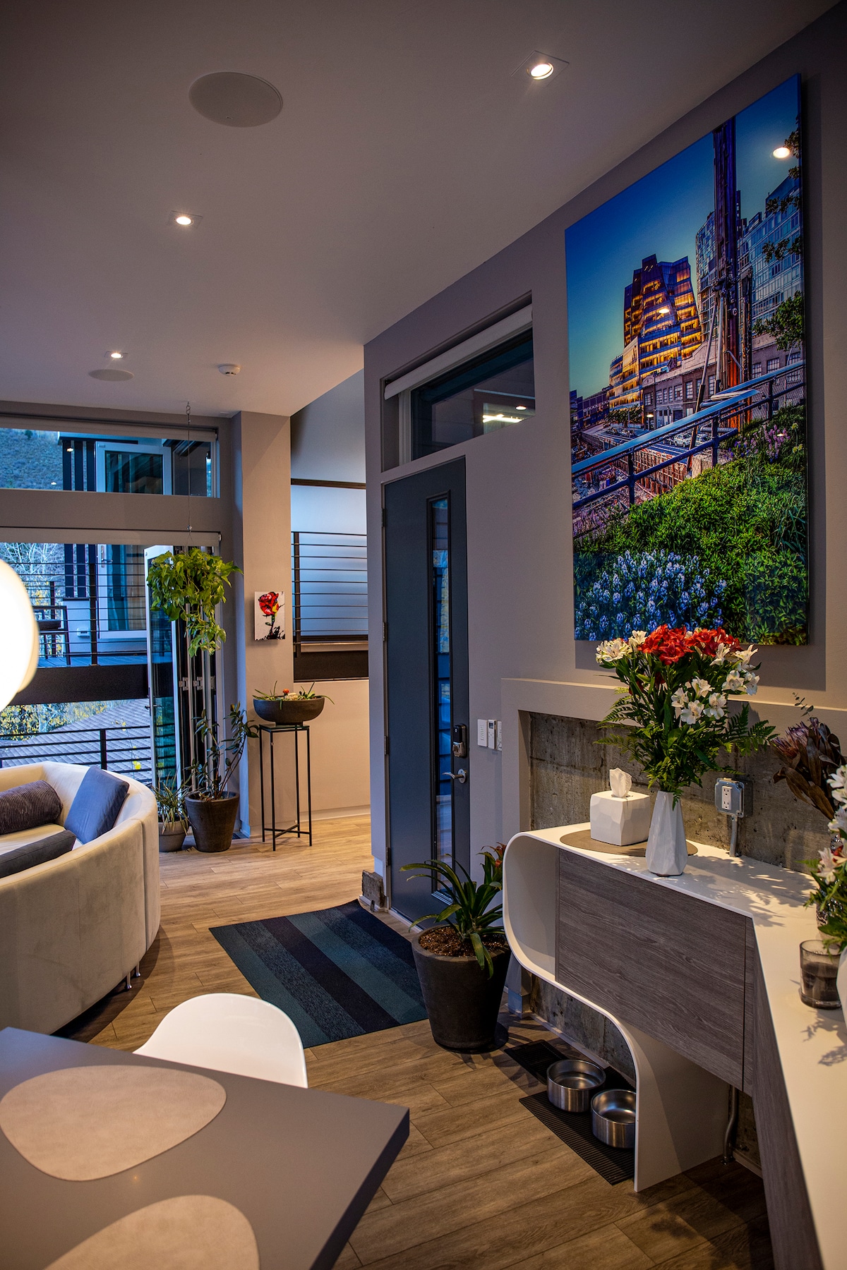 An entryway features modern decor, including a large artwork against the wall depicting a city skyline. A side table with flowers and plants adds a touch of greenery, while the open space is illuminated by recessed lighting and natural light from nearby windows.