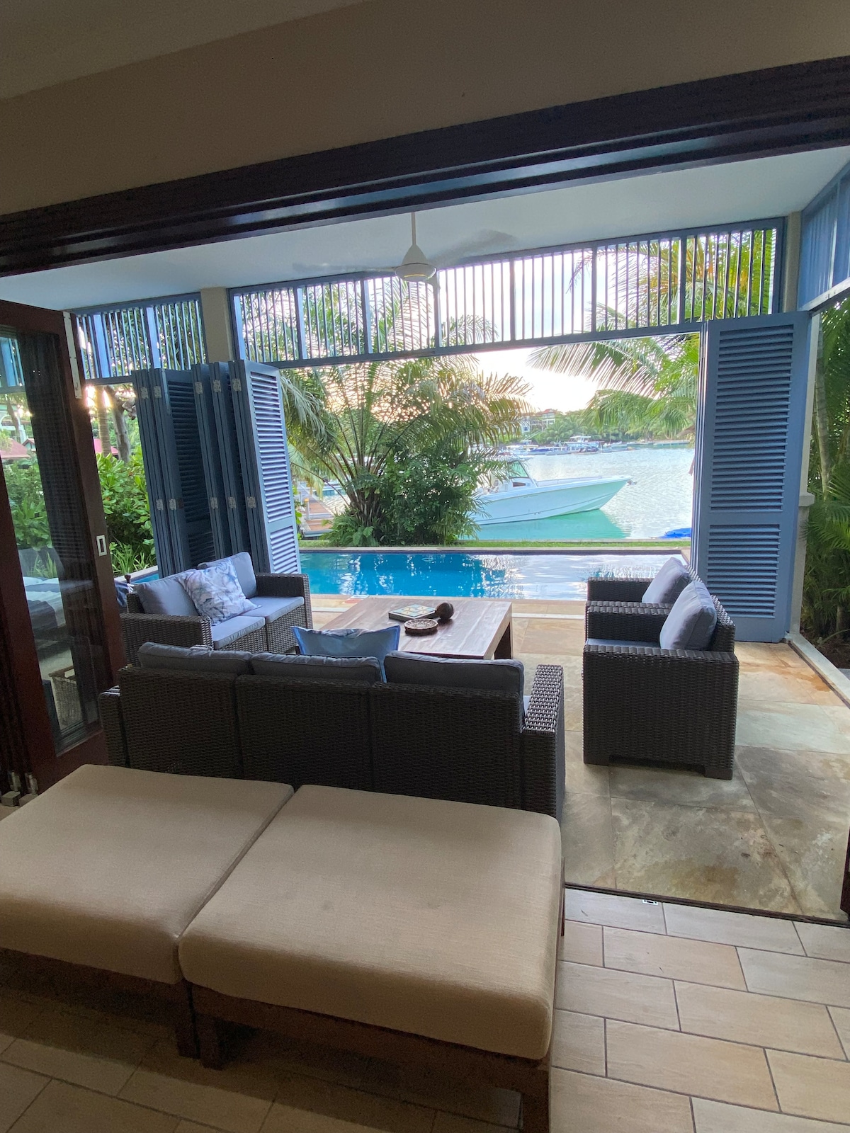 An outdoor seating area is set up with comfortable wicker furniture and cushions, overlooking a private pool and tropical garden. Lush greenery frames the view of the marina, creating a serene atmosphere. Large sliding doors provide seamless access to the indoor space.