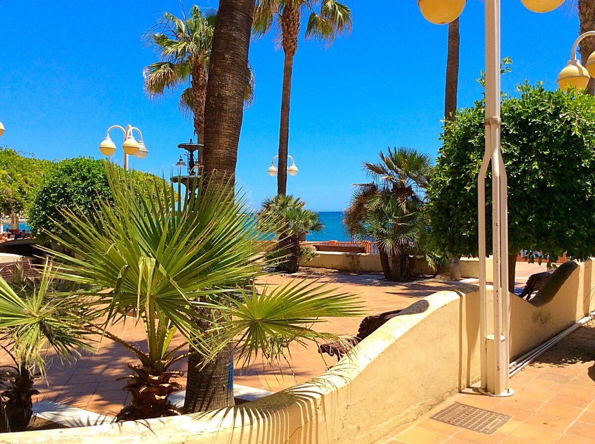 A vibrant outdoor space is presented, featuring palm trees and lush greenery. The sun-drenched walkway leads to a glimpse of the ocean, with the blue sky overhead. Ambient light fixtures are visible, adding to the inviting atmosphere of the coastal setting.
