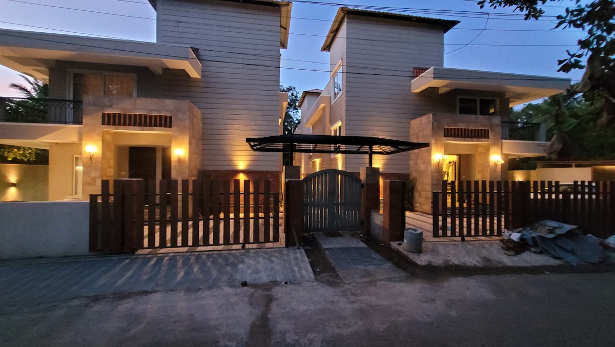 Two modern villa buildings are shown at twilight, featuring a welcoming exterior with well-lit stone accents. A wooden fence surrounds the entrance, with a metal gate positioned at the center. Warm light emanates from fixtures above, highlighting the inviting ambiance.