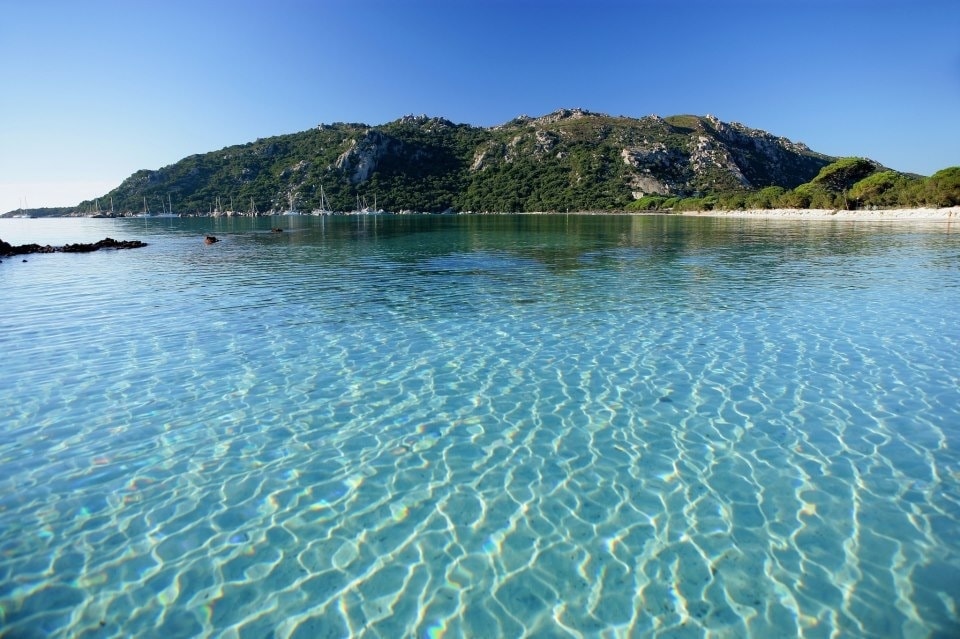 A serene view of clear blue water reflects the sky, creating a calm atmosphere. Gentle ripples can be seen on the surface, with the distant outline of lush green hills and rocky formations framing the scene. The tranquility invites relaxation and appreciation of nature.