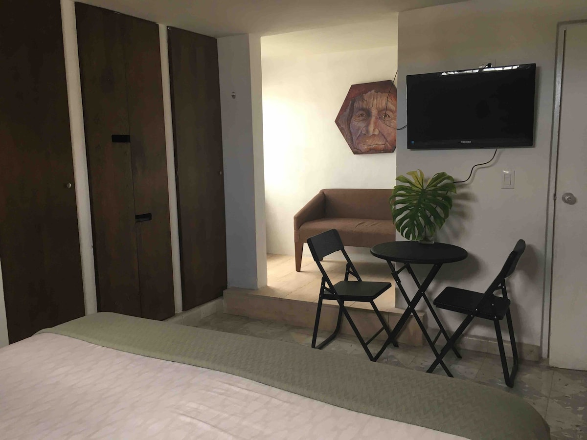 A studio space is shown with a spacious bed in the foreground. Two wooden closets are visible along the wall, and a flat-screen TV is mounted nearby. A small round table and two folding chairs create a cozy sitting area next to a sofa, enhancing the functional layout.