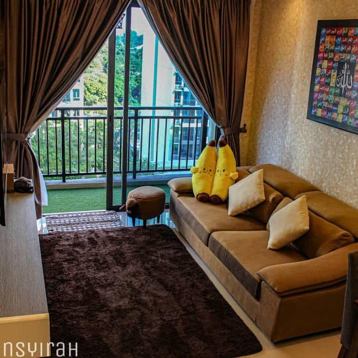 A cozy living area is furnished with a beige sectional sofa and two plush cushions. Large windows draped in dark curtains offer a view of the outdoor greenery. A small rug lies on the glossy floor, while decorative plush toys add a playful touch to the space.