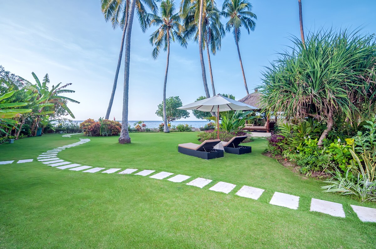 A well-maintained garden features a winding stone pathway leading to the ocean. Palm trees provide shade over lounge chairs next to a large parasol. Lush greenery and tropical plants create a serene setting, enhancing the outdoor space's connection to nature.