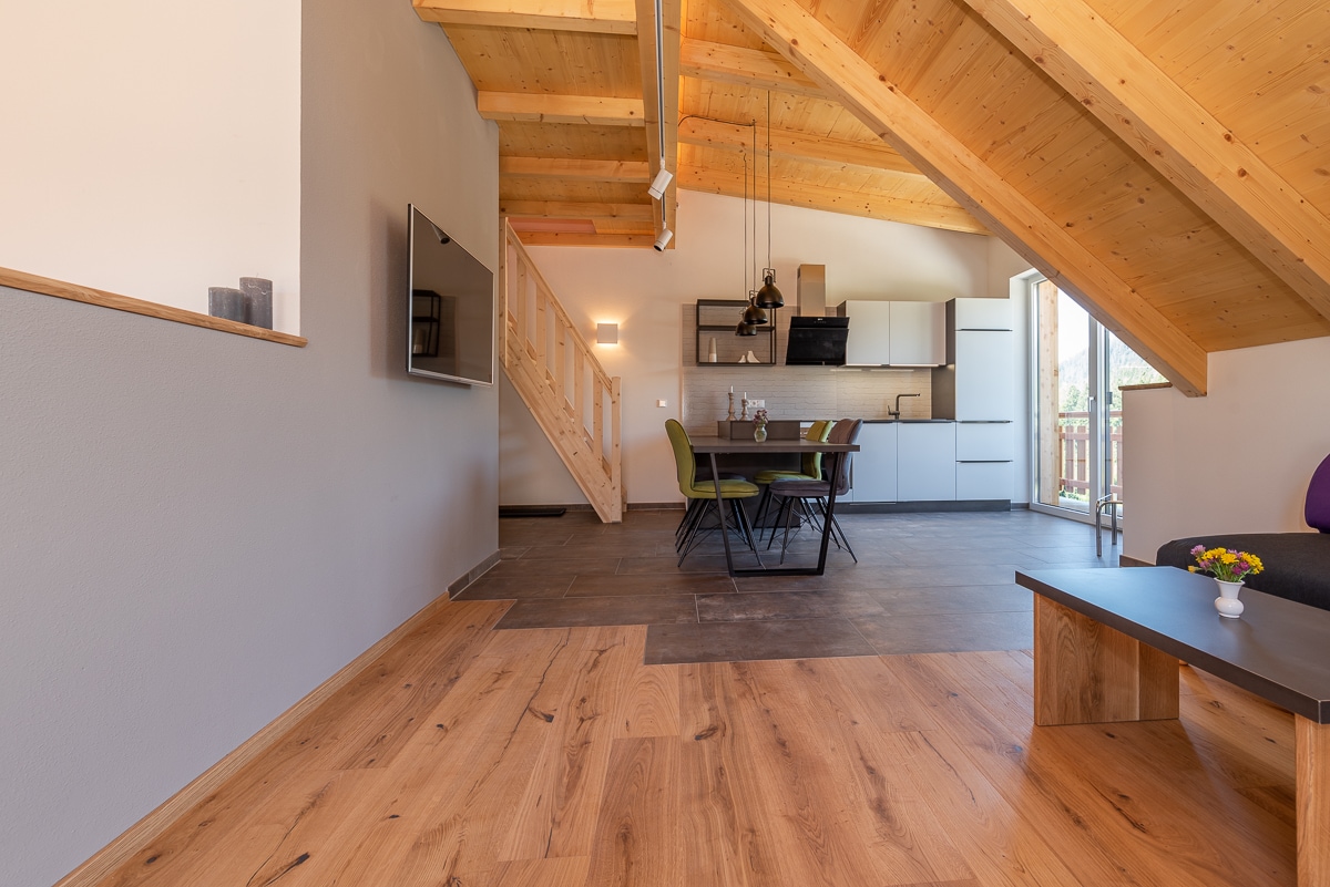 An open living space is showcased with wooden beams accentuating the high ceiling. A dining table with four chairs is positioned near a modern kitchen. A flat-screen TV is mounted on the wall, and large glass doors lead to a balcony, allowing natural light to enter.