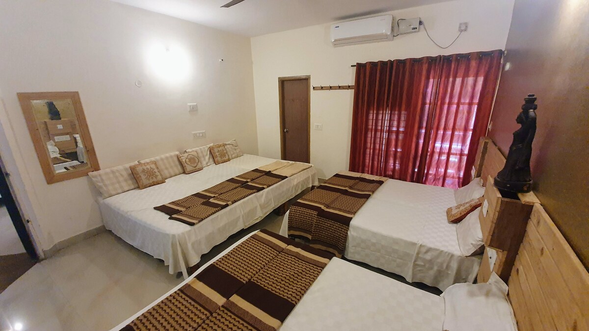 The room features three king cots and one single cot, arranged with neutral bedding. Natural light filters through a sheer curtain, and a wall-mounted air conditioning unit is visible. A wall mirror and a door lead to the attached bathroom, enhancing the room's functionality.