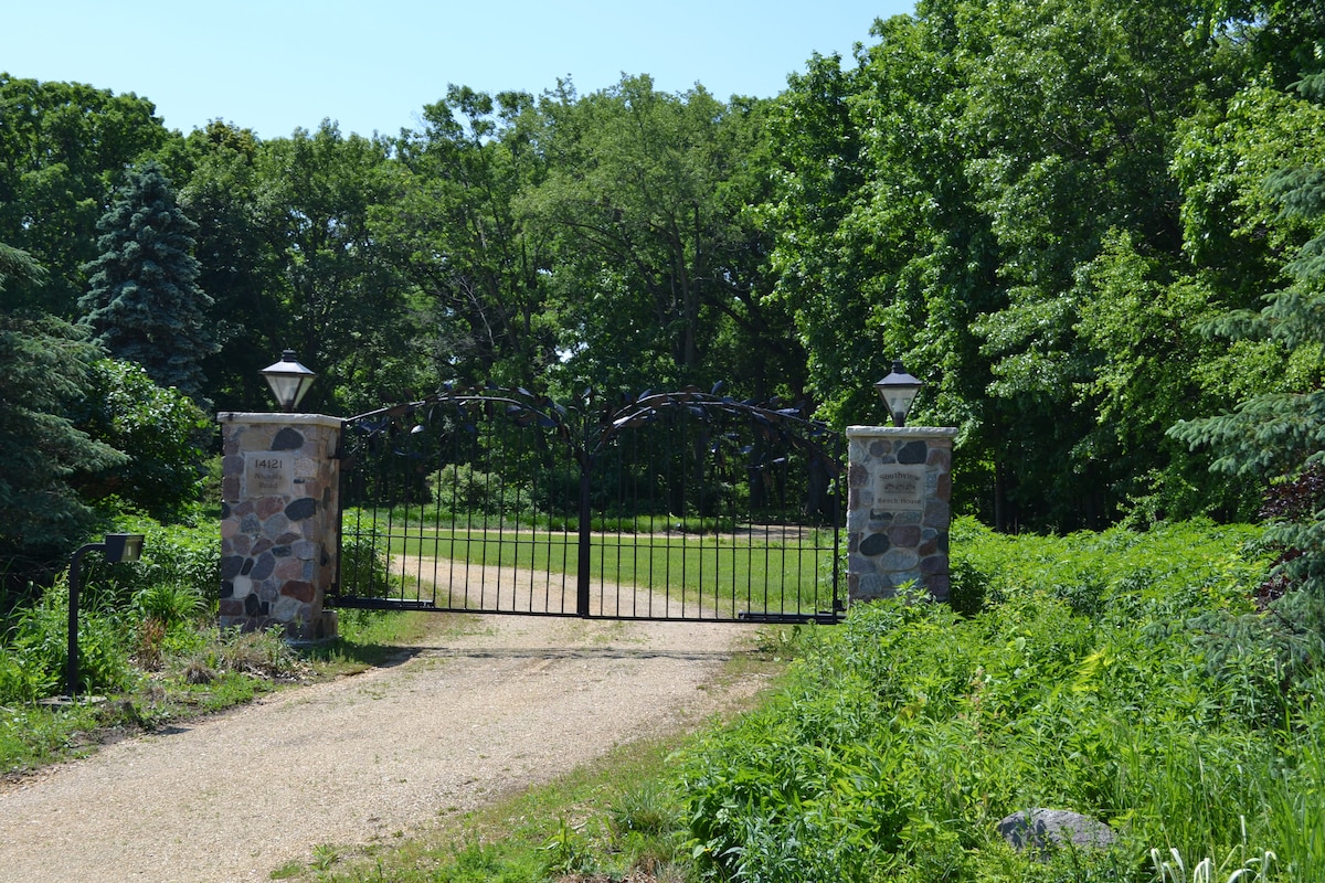 A gated entrance welcomes guests to the property, featuring stone pillars with decorative lighting. Lush greenery surrounds the gravel driveway, leading to expansive grassy areas beyond the gate. Mature trees add depth to the landscape, enhancing the natural setting.