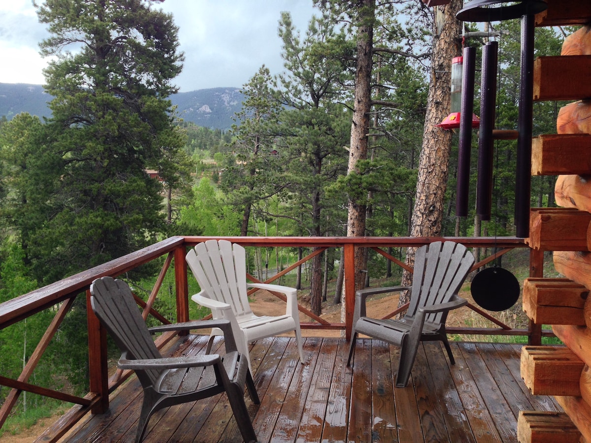 A deck area features four Adirondack chairs arranged comfortably, inviting relaxation with natural surroundings. Tall trees and rolling hills are visible in the background, enhancing the serene atmosphere. A wind chime hangs nearby, adding a gentle visual element to the outdoor space.