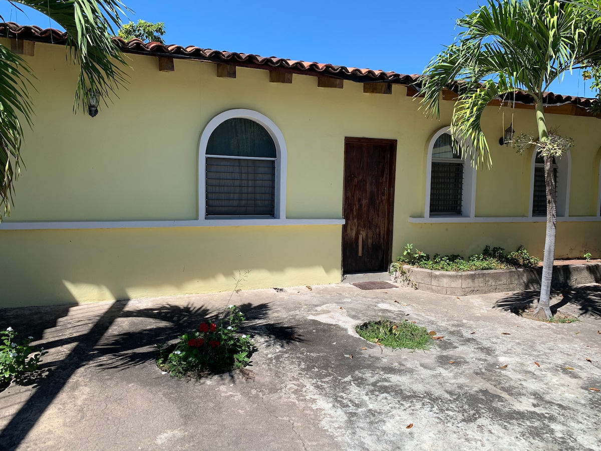 A single-story exterior of a yellow house is presented, featuring arched windows and a wooden door. Lush green plants and flowers are visible in the foreground. The property is set against a clear blue sky, emphasizing a calm and inviting environment.