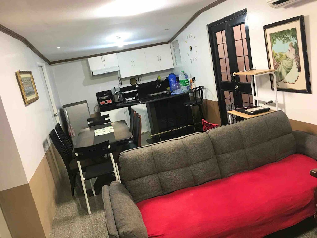A living area features a gray sofa with a red throw blanket, providing a cozy seating option. A dining table with black chairs is positioned near the kitchen area. Clean white cabinetry and dark countertops are visible in the background, contributing to a functional space.