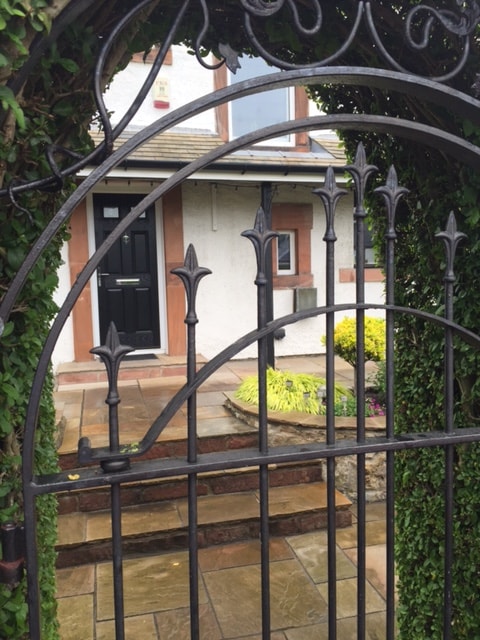 An ornate wrought iron gate is framed by lush greenery, leading to a neatly paved pathway. Visible steps ascend towards a welcoming entrance, featuring a dark door with contemporary hardware. Colorful plants are positioned nearby, complementing the serene garden ambiance.
