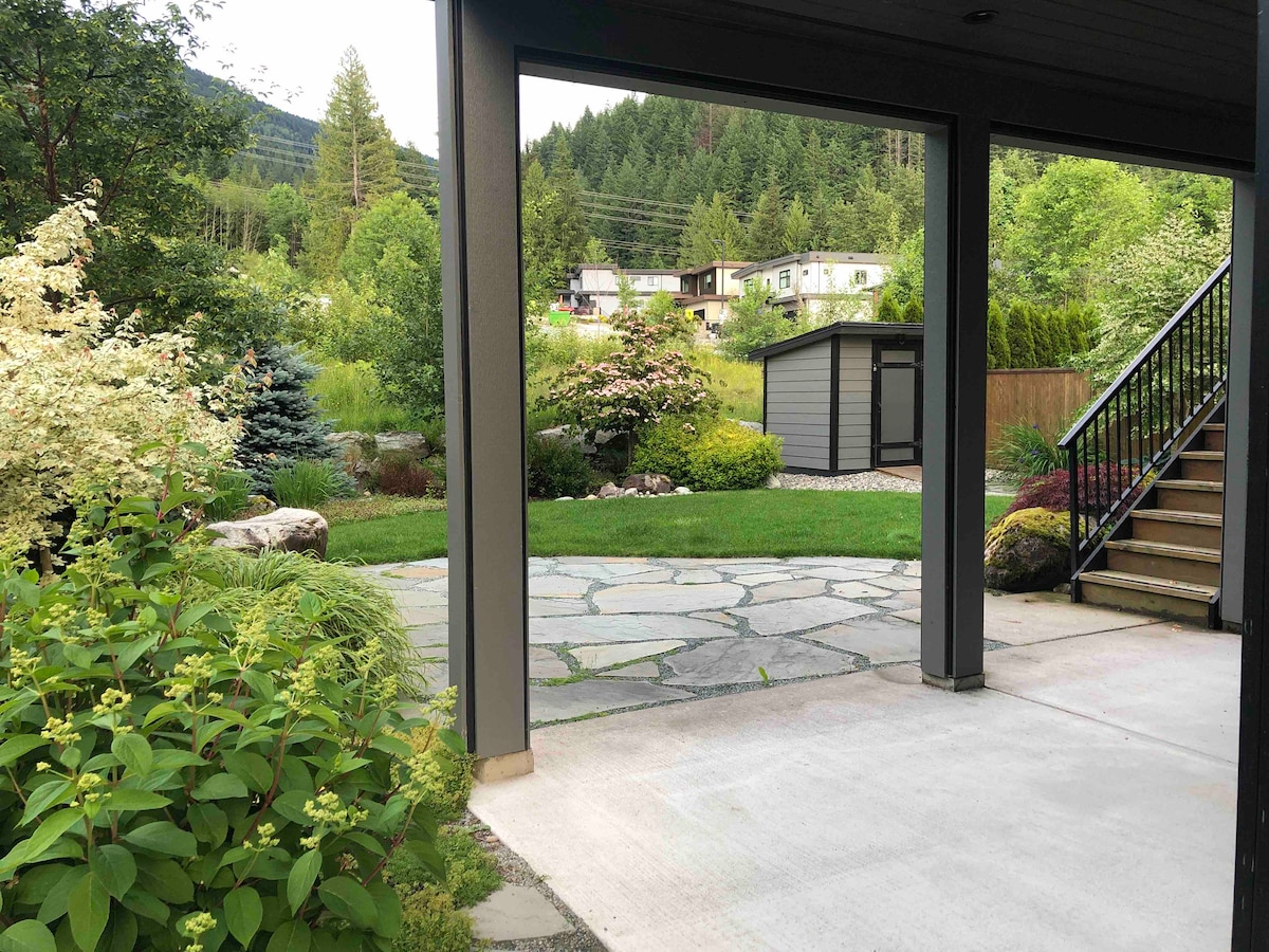 An inviting outdoor area is framed by structural columns, showcasing a landscaped garden with various plants and trees. A stone pathway leads to a grassy space and a nearby shed, while the backdrop features rolling hills and residential homes nestled among greenery.