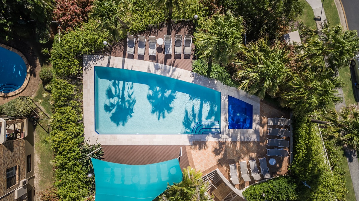 An aerial view captures a large swimming pool surrounded by tropical greenery. Sun loungers are arranged neatly along the pool's edge, while a smaller hot tub is positioned nearby. The vibrant blue water reflects the clear sky and palm shadows.