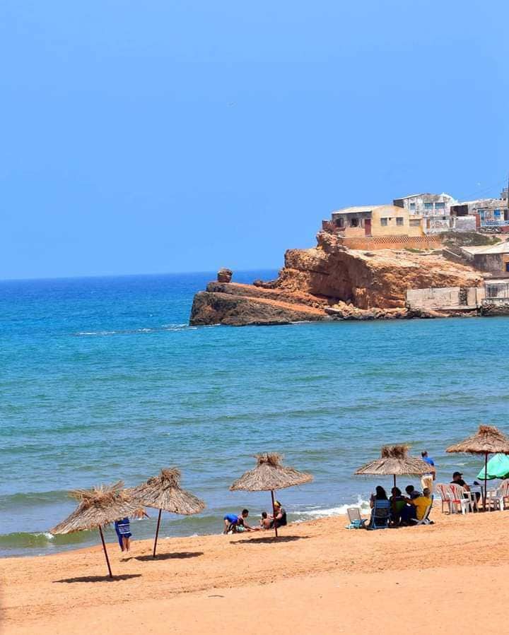 The image features a sandy beach with thatched umbrellas providing shade. In the background, a rocky coastline is visible, with a cluster of houses perched above the water. The ocean displays shades of blue, with gentle waves lapping at the shore.