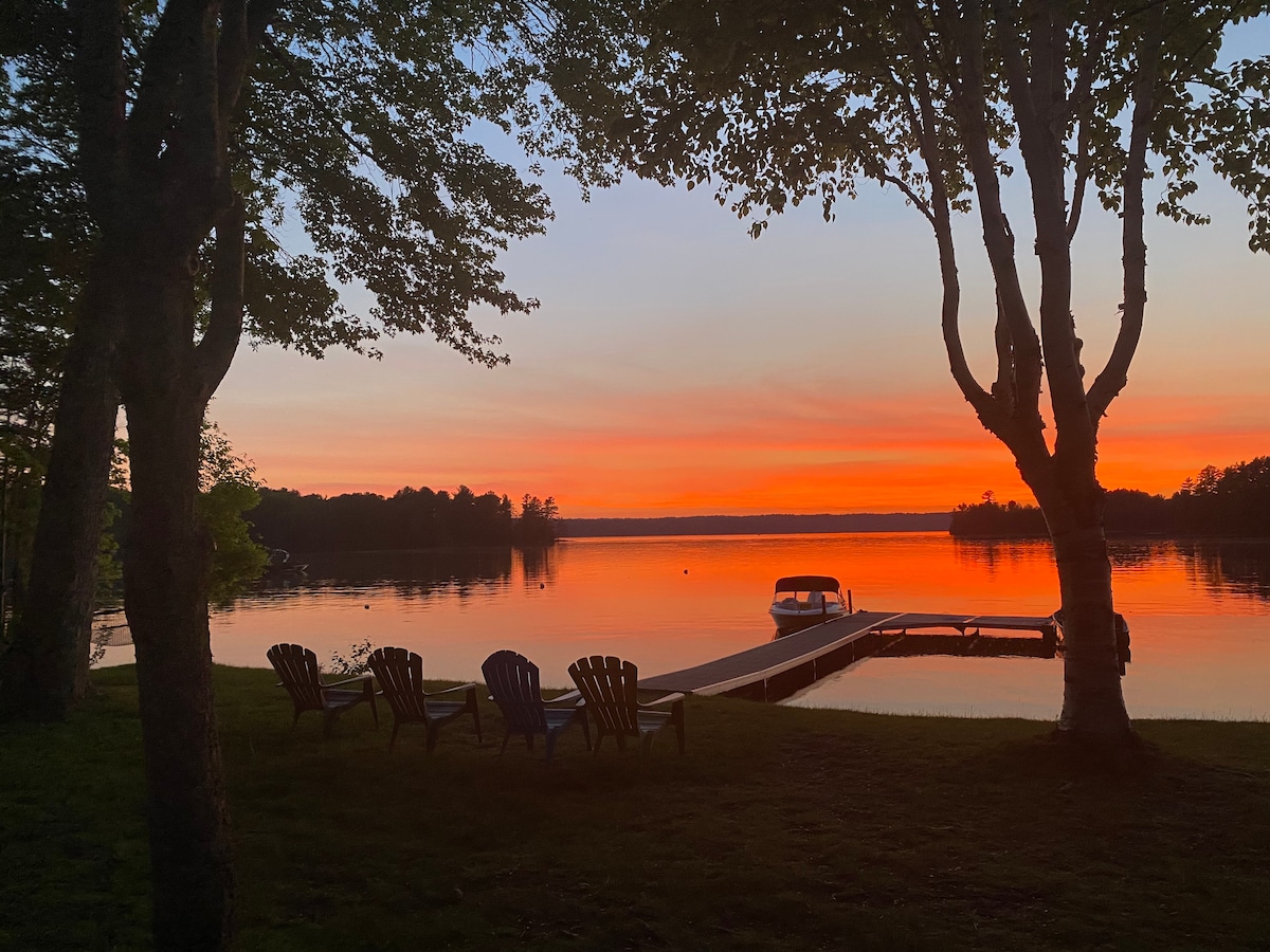 A serene lake view is captured at sunset, featuring vibrant hues of orange and pink reflected in calm waters. Two trees frame the scene, which includes a boat dock and several adirondack chairs arranged on a grassy area, inviting relaxation by the water.