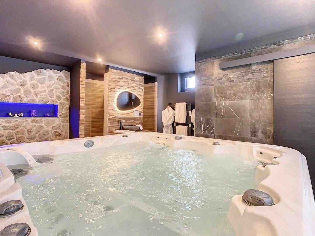 A spacious Jacuzzi designed to accommodate five people is featured, surrounded by elegant stone walls and modern lighting. Soft reflections are visible on the water surface, and a plush bathrobe is seen nearby, suggesting a comfortable and relaxed environment.