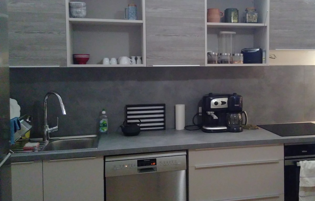 The kitchen features modern cabinetry and a sleek countertop. A stainless steel sink is positioned beside a dishwasher and an array of kitchen appliances including a coffee maker. Shelves above display various containers and dishware, contributing to a functional cooking space.