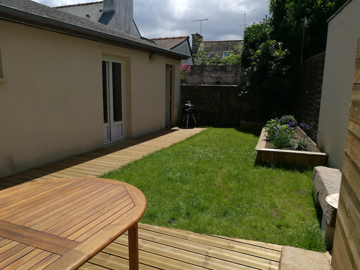 A spacious garden area features a wooden deck and a well-maintained lawn. The sunlight brightens the green space, complemented by flowering plants in raised beds. A charcoal grill is positioned against the wall, and a set of wooden outdoor furniture is present for relaxation.
