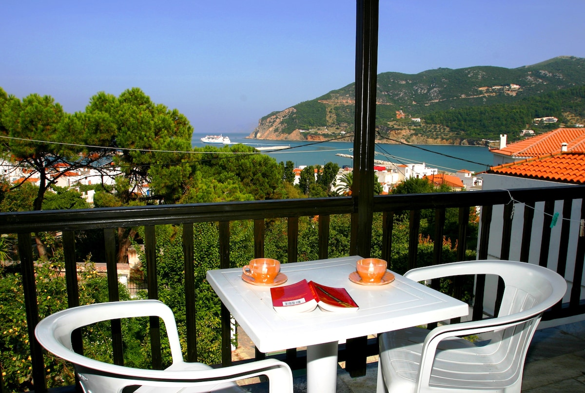 A private balcony is shown, featuring a small white table set for two with orange cups, overlooking a scenic ocean view. The coastline and hills are visible in the background, framed by greenery and rooftops of neighboring buildings.