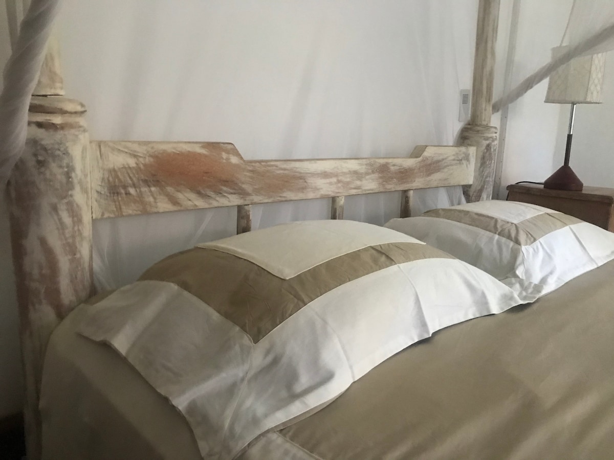A spacious bed features a unique wooden headboard with a natural finish. The bed is adorned with soft, cream-colored linens and two neatly arranged pillows. Gentle light from a bedside lamp adds warmth to the room.