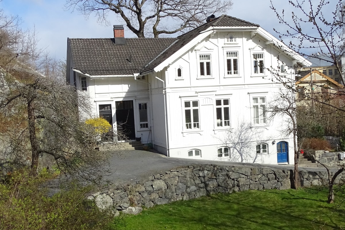 The charming white wooden building showcases traditional Norwegian architecture with large windows and a well-maintained garden. A stone wall surrounds the property, and a pathway leads to the entrance. The structure is set against a backdrop of greenery and a clear blue sky.