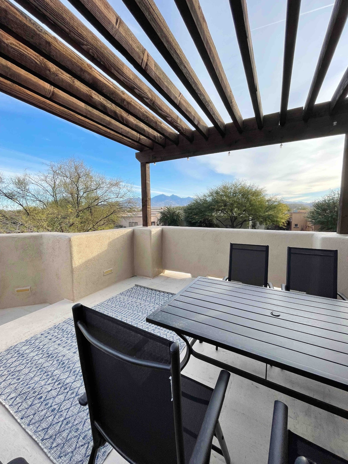 An outdoor sky deck features a shaded area with wooden beams overhead. A rectangular table with dark chairs is set on a light-colored surface, accompanied by a patterned rug. Scenic views of trees and distant mountains can be seen in the background.
