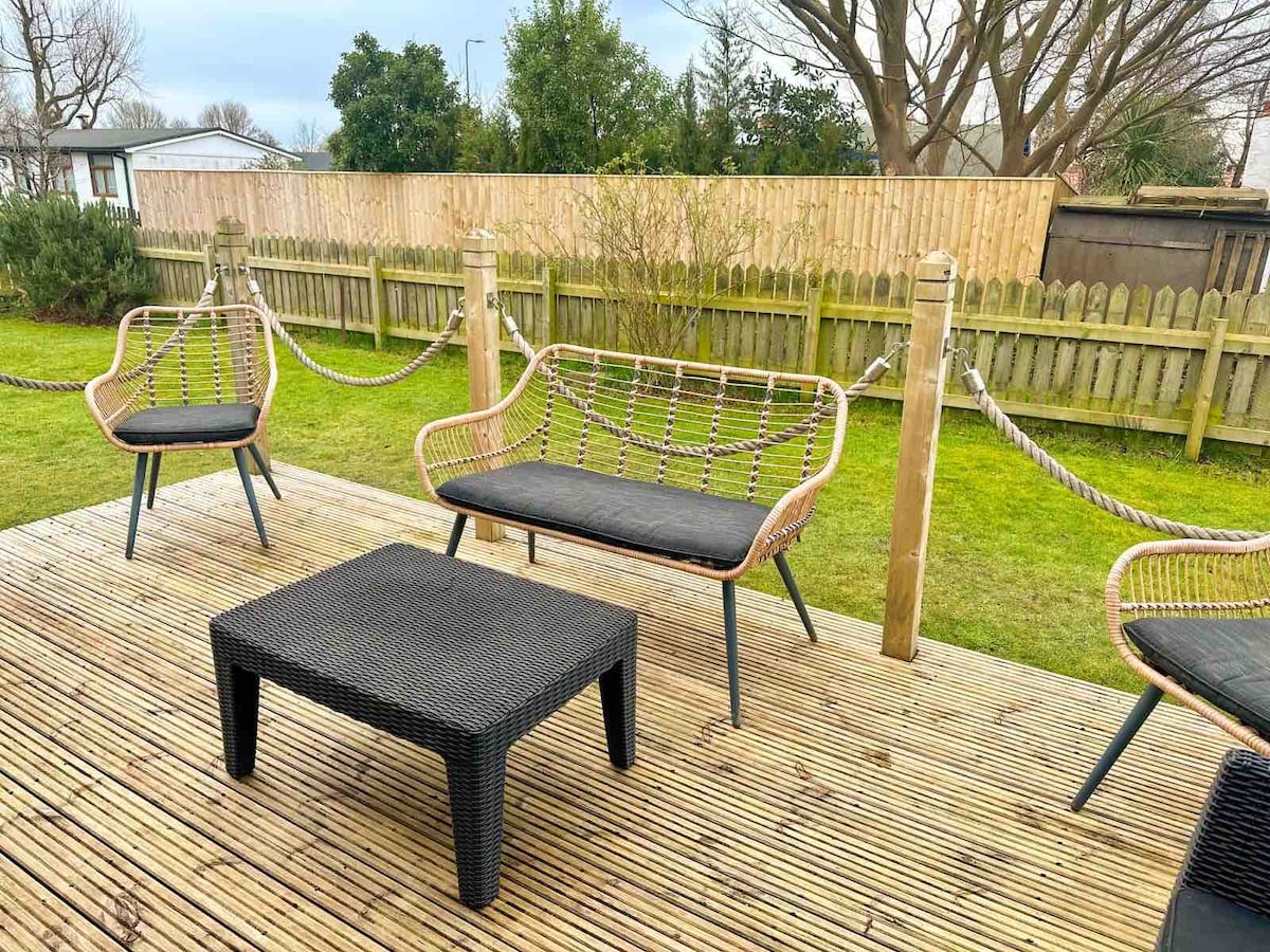 A wooden deck area features four stylish chairs with black cushions and a low black coffee table. A surrounding fence and greenery provide a sense of privacy and connection to nature, enhancing the outdoor space's appeal.