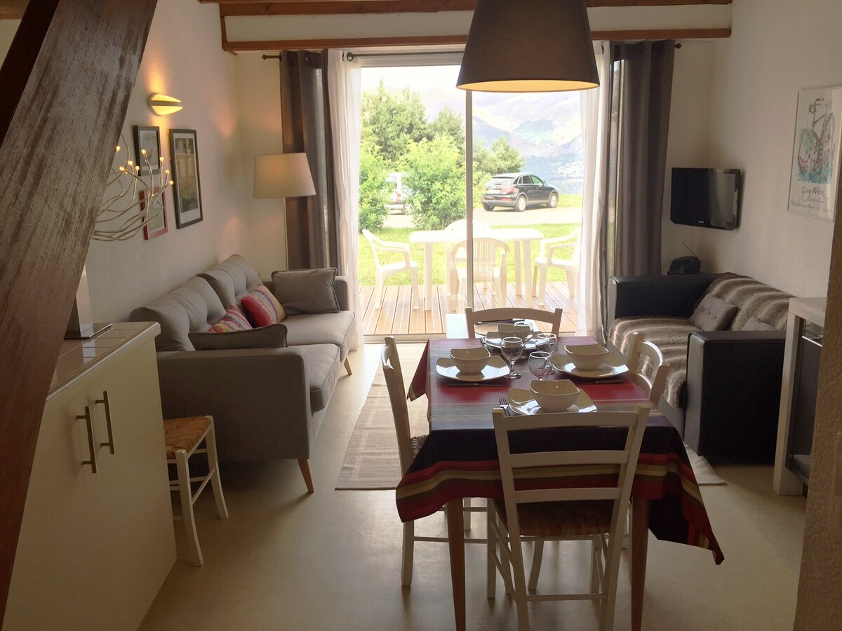 The spacious living area features comfortable seating with two sofas and a dining table set for six. Large glass doors open to a wooden terrace with a view of the outdoors. A flat-screen TV is mounted on the wall for entertainment.